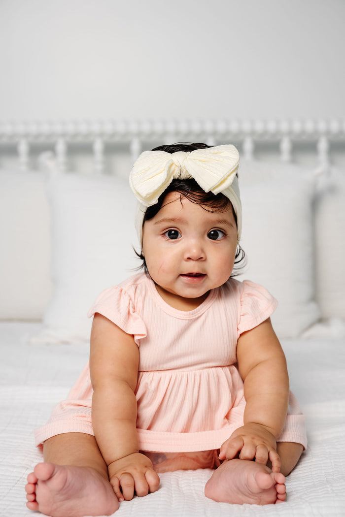 Baby wearing a pink dress and white headband with a bow, sitting on a white surface.