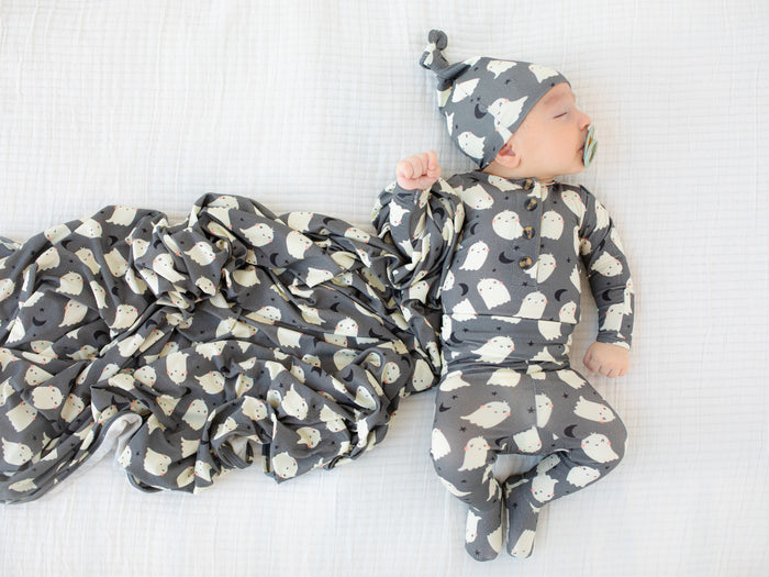 Baby in a gray and white patterned onesie and hat lying on a white blanket.