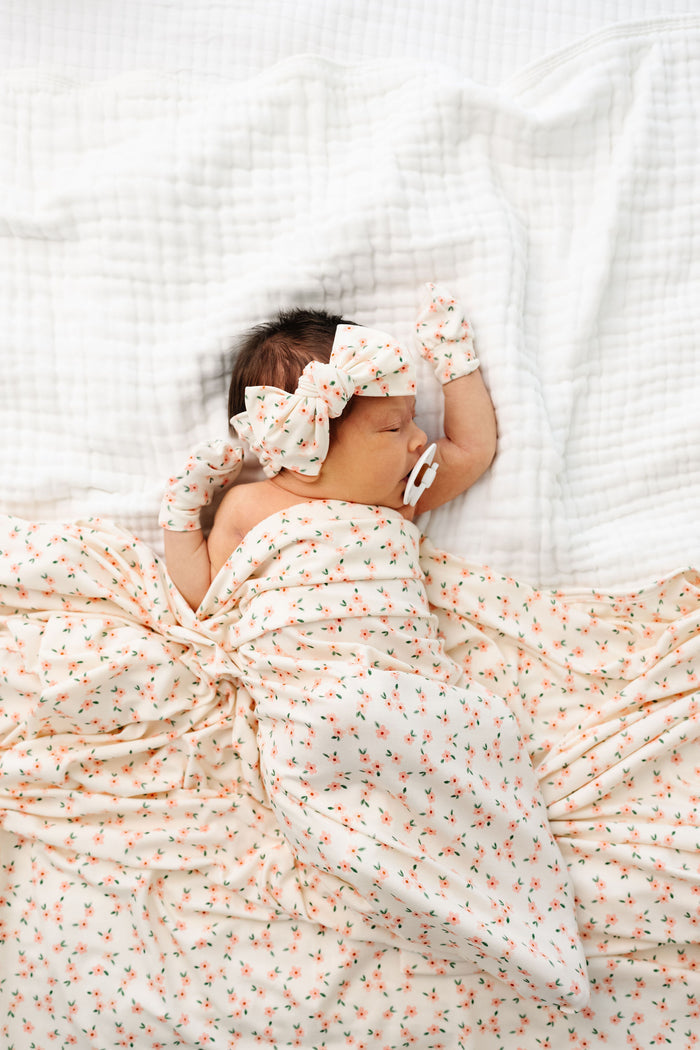 lottie headband bundle newborn girl laying on bed swaddle blanket, knit headband, and no scratch mittens.